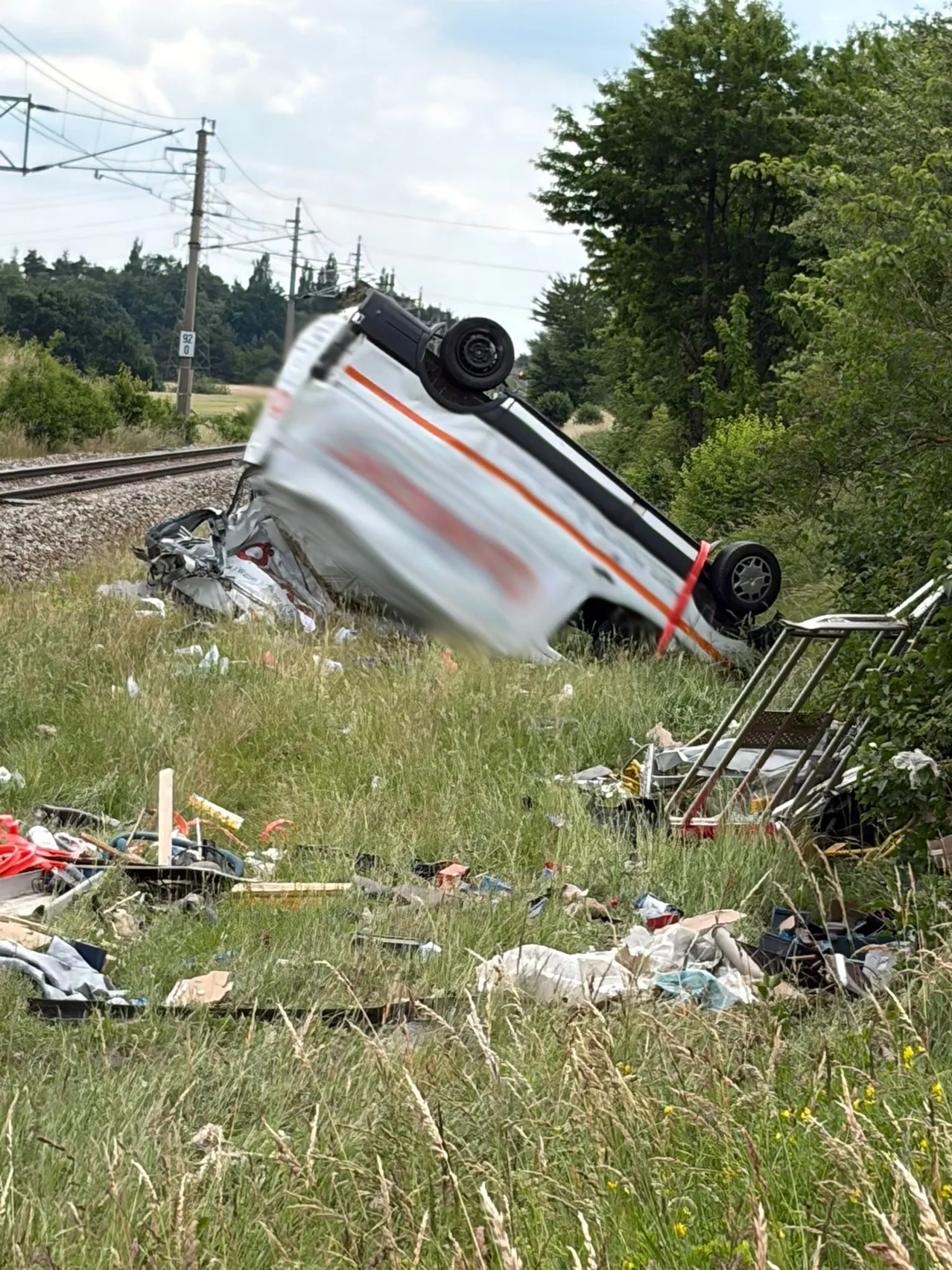 Ein umgestürztes Fahrzeug liegt auf seiner Seite in einem Grasfeld in der Nähe einer Bahnstrecke, mit verstreutem Schutt drumherum.