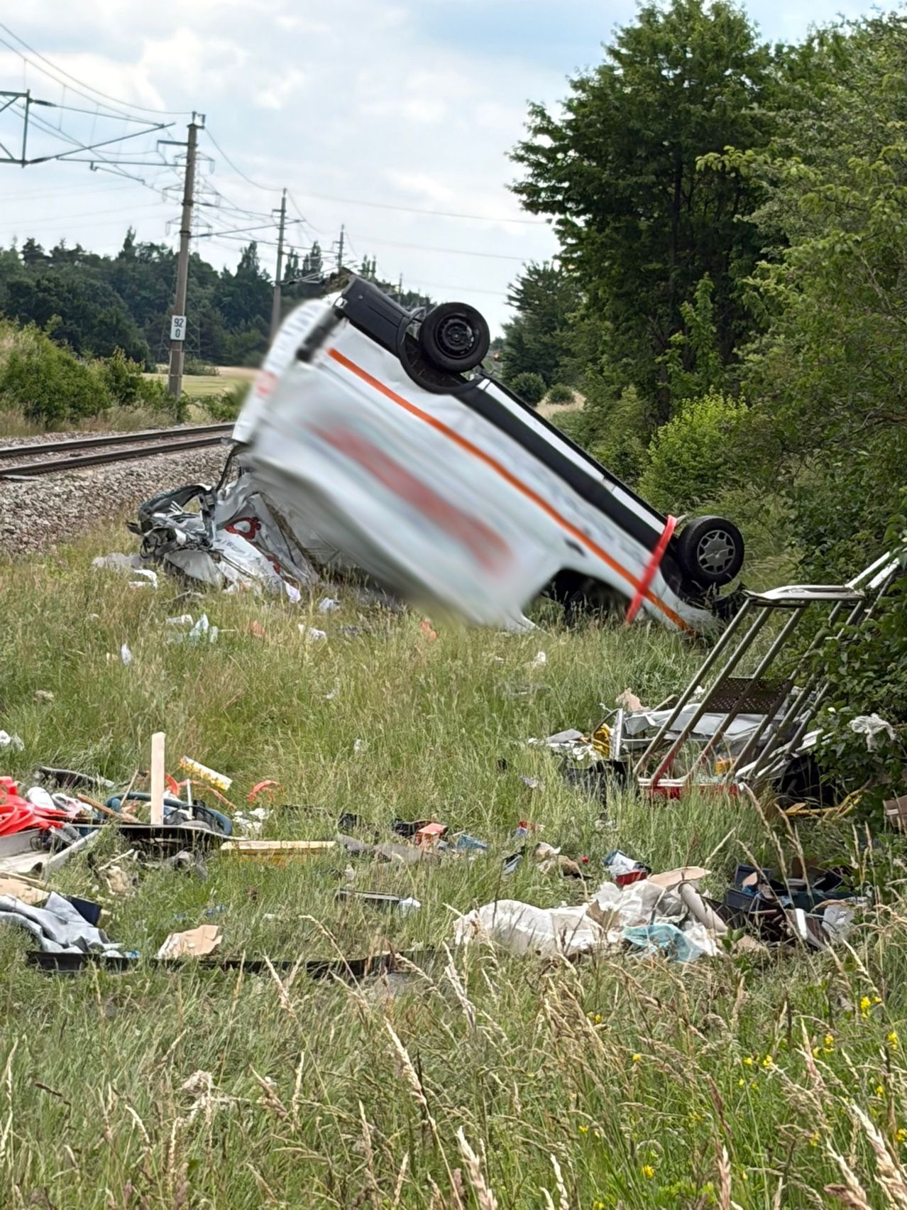 Ein umgestürztes Fahrzeug liegt auf seiner Seite in einem Grasfeld in der Nähe einer Bahnstrecke, mit verstreutem Schutt drumherum.