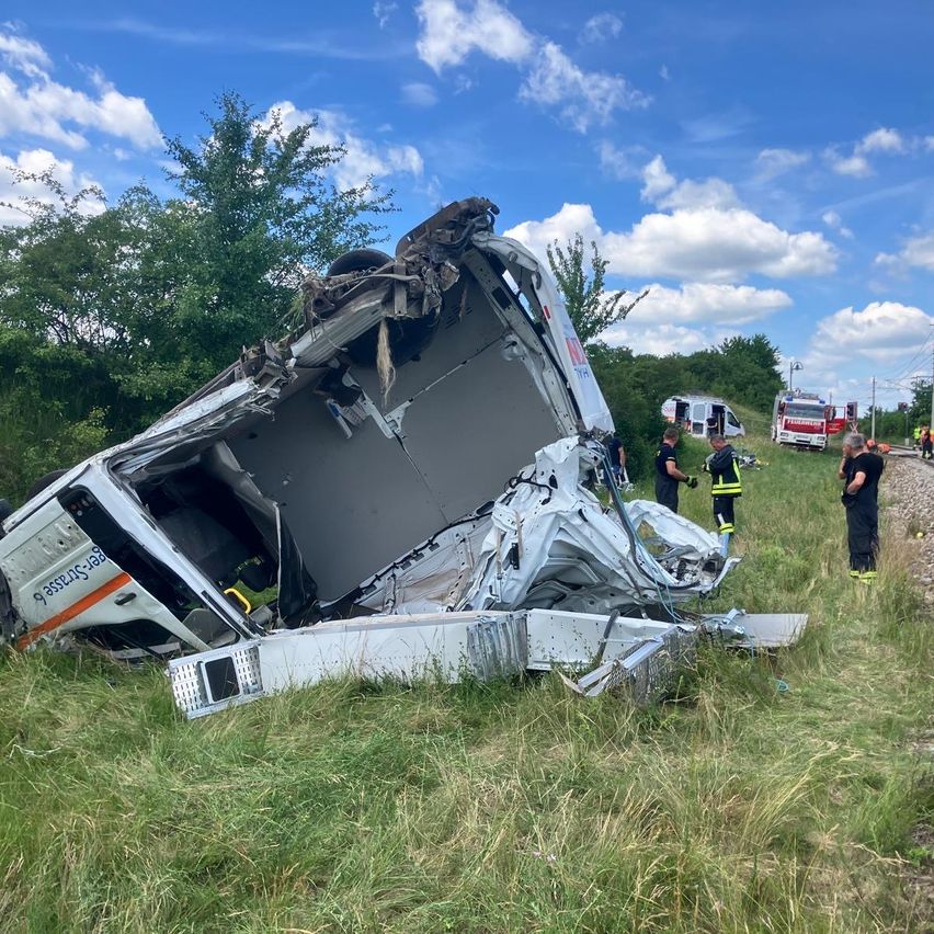 Ein demoliertes Fahrzeug liegt auf der Seite in einem Grasfeld. Rettungskräfte sind in der Nähe, und ein Feuerwehrwagen ist im Hintergrund geparkt.