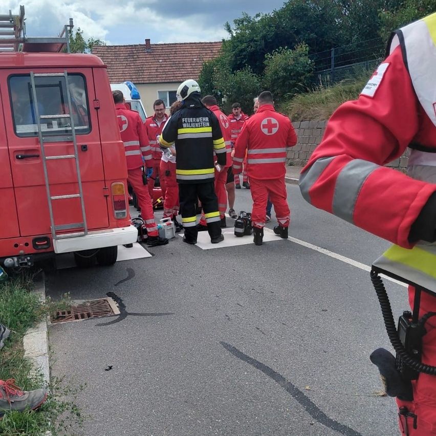 Feuerwehrleute in roten Uniformen, einige mit Helmen, kümmern sich um einen Vorfall auf der Straße. Ein roter Feuerwehrwagen steht in der Nähe.