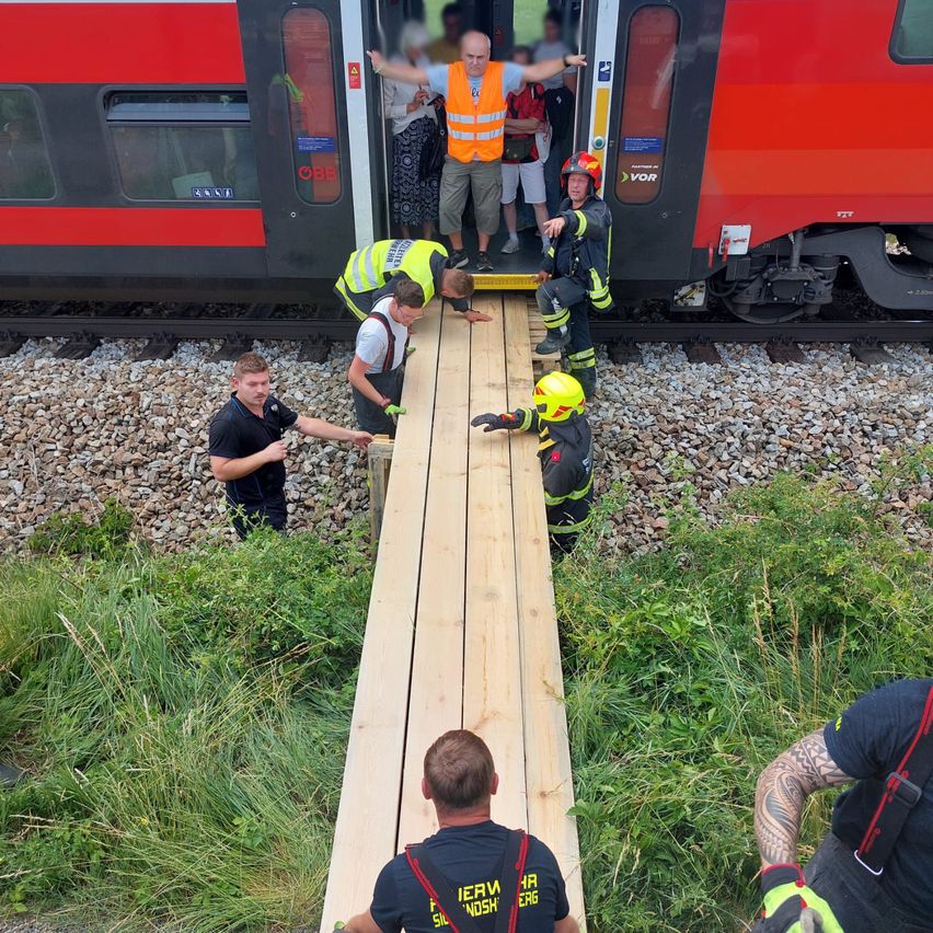Feuerwehrleute helfen Passagieren beim Verlassen eines entgleisten Zuges mit einer Rampe. Sie stehen auf Kies mit dem Zug auf den Gleisen hinter ihnen.