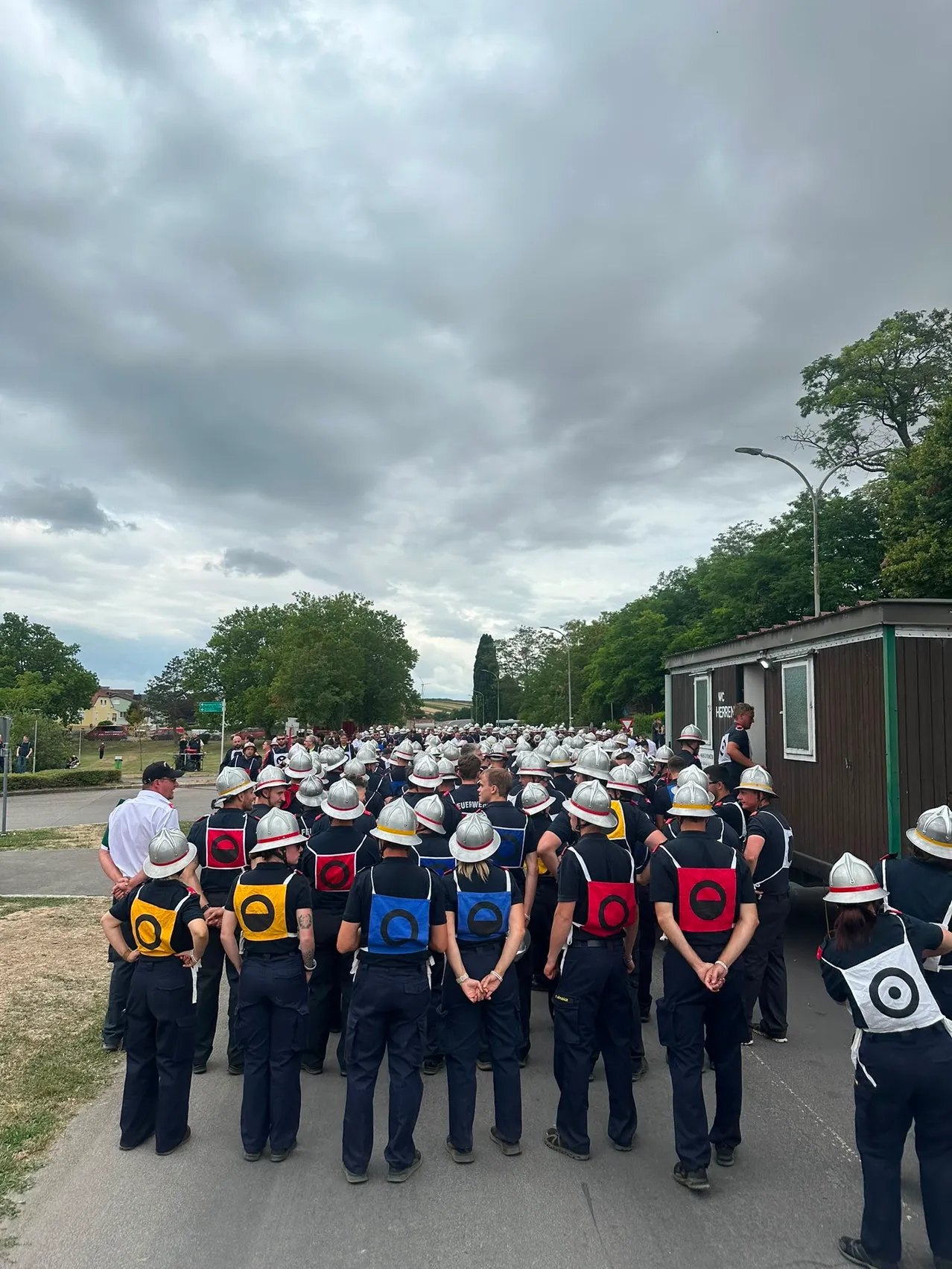 Eine Gruppe von Personen in Uniformen und Helmen steht im Freien in einer Reihe, mit einem Gebäude und Bäumen im Hintergrund.
