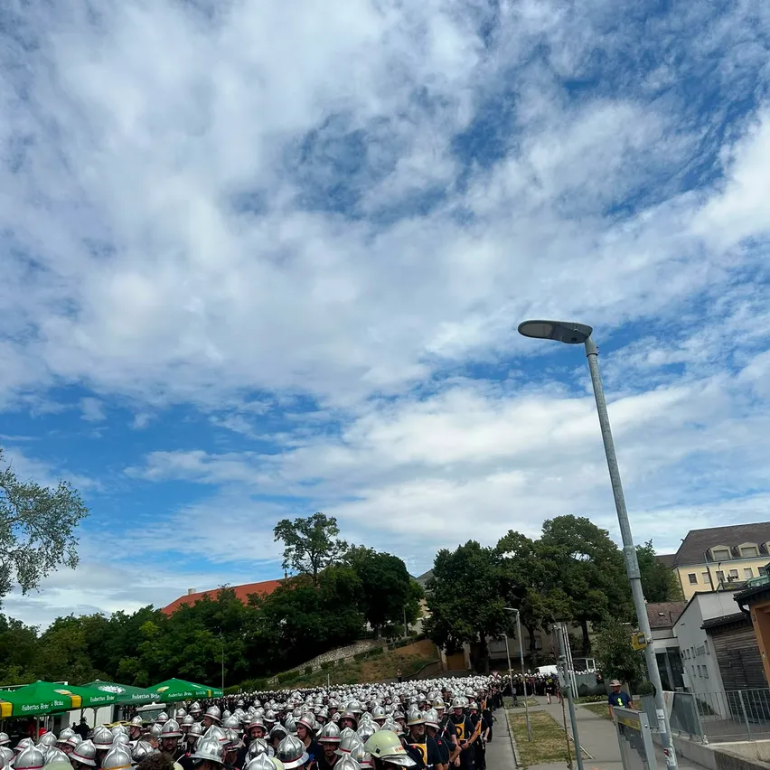 Eine große Gruppe von Menschen in Helmen und Uniformen marschiert auf einer Straße mit Bäumen und Gebäuden im Hintergrund.
