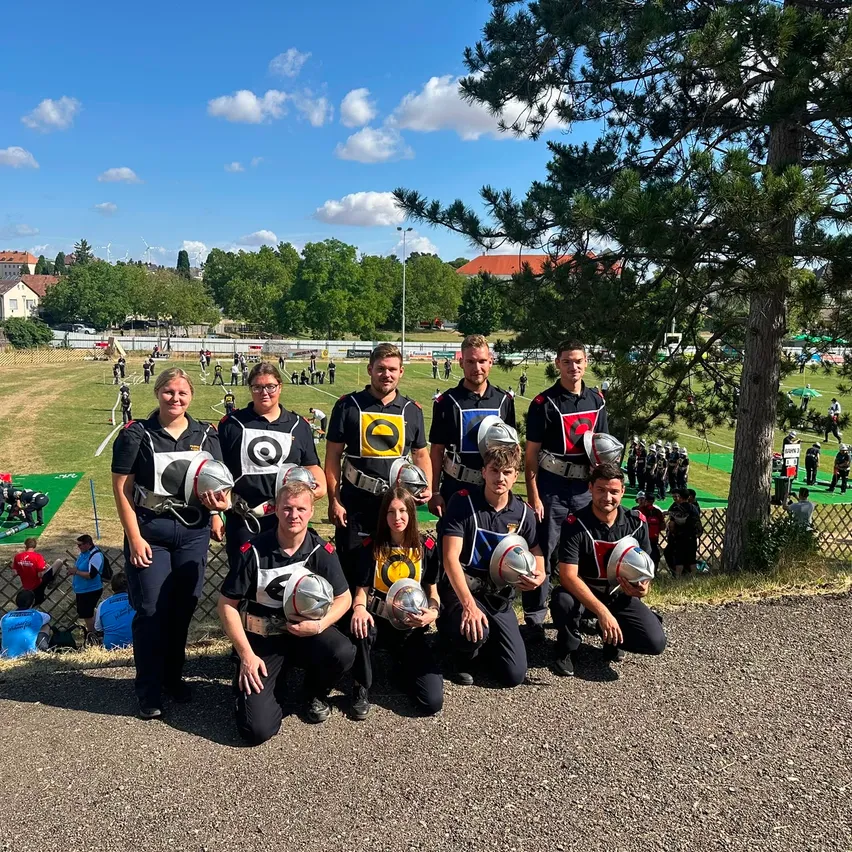 Eine Gruppe von Menschen, möglicherweise ein Sportteam, posiert für ein Foto, während sie Uniformen und Helme tragen. Im Hintergrund sieht man ein Grasfeld mit einigen Personen und einem Baum.
