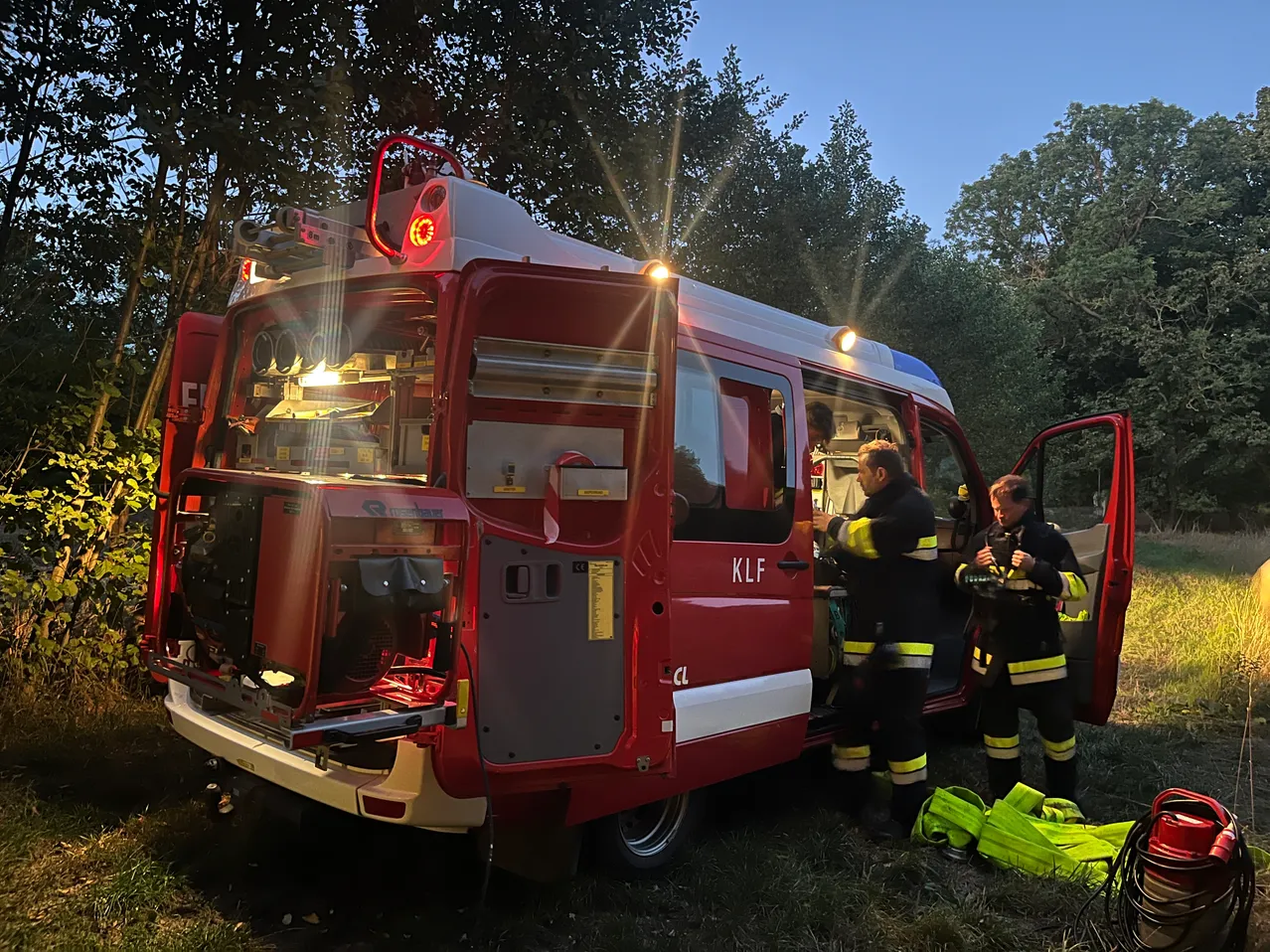 Zwei Feuerwehrleute stehen vor einem roten Feuerwehrwagen mit offenen Hecktüren, einer im Fahrzeug und der andere mit Ausrüstung. Der Wagen steht auf einer Wiese mit Bäumen im Hintergrund.