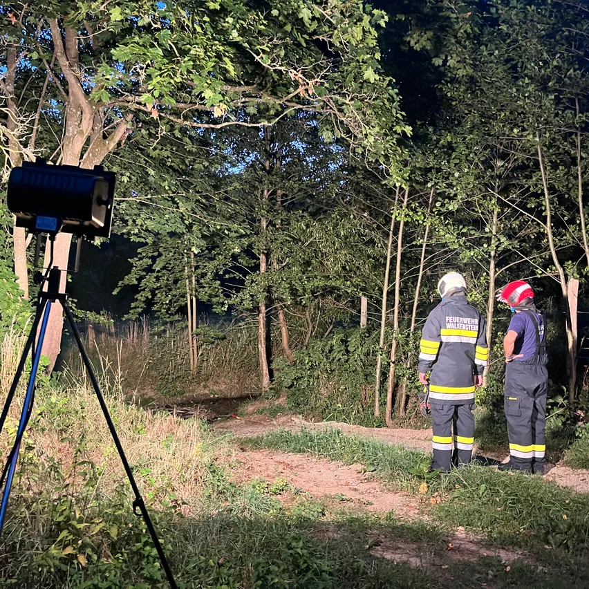 Zwei Feuerwehrleute in Uniform stehen auf einem Feldweg in einem bewaldeten Gebiet, einer trägt einen Helm. Eine Kamera auf einem Stativ ist links positioniert.