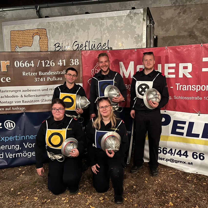 Fünf Personen posieren für ein Foto vor einem Banner. Sie tragen alle Helme und Uniformen.