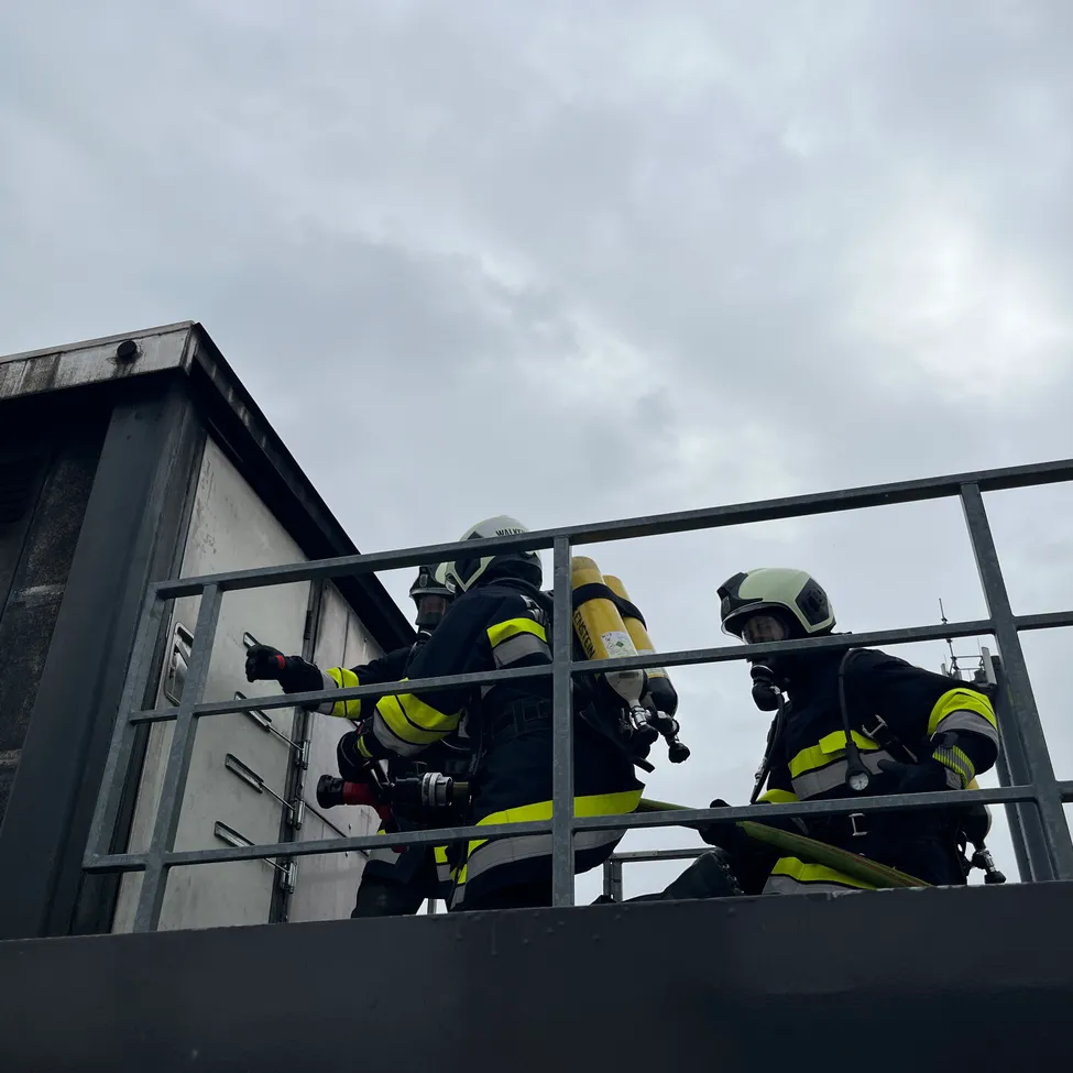 Drei Feuerwehrleute in gelben und schwarzen Uniformen sind auf dem Dach eines Gebäudes. Sie sind mit Helmen, Sauerstoffflaschen und Feuerlöschern ausgestattet.