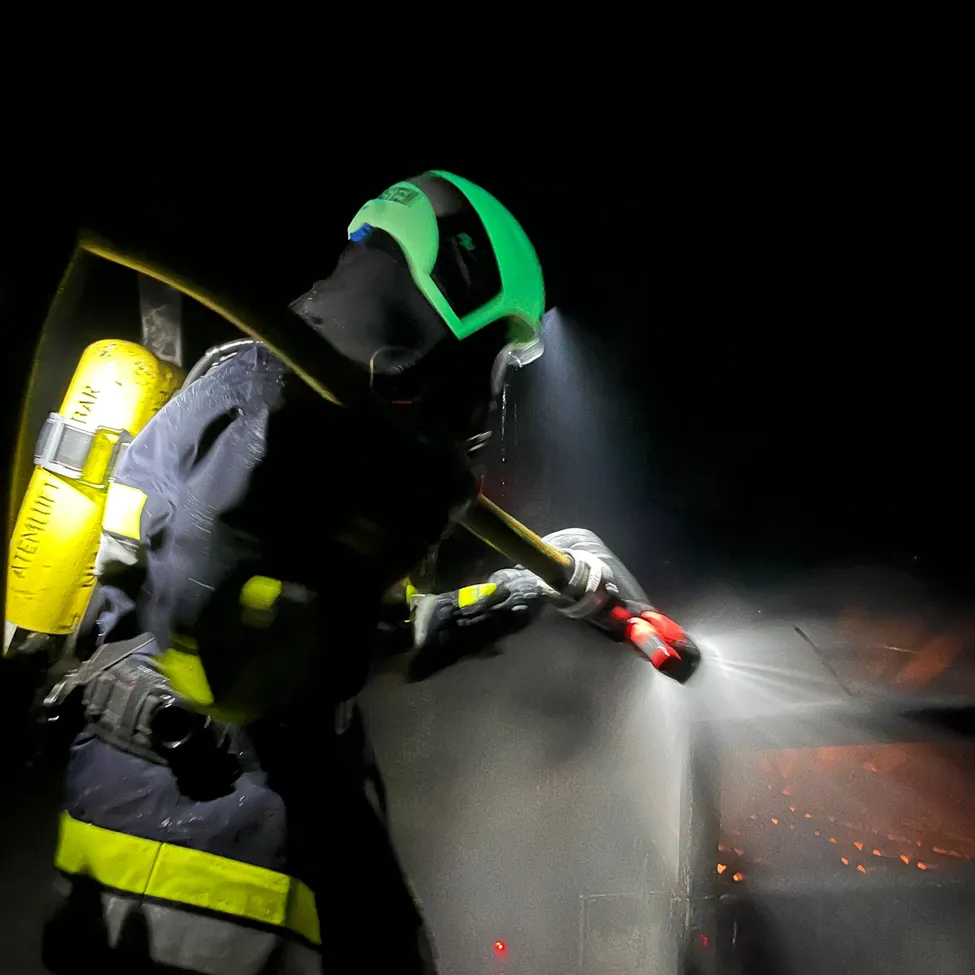 Ein Feuerwehrmann in einem grünen Helm und einem gelben und schwarzen Uniform spritzt Wasser aus einem Schlauch.