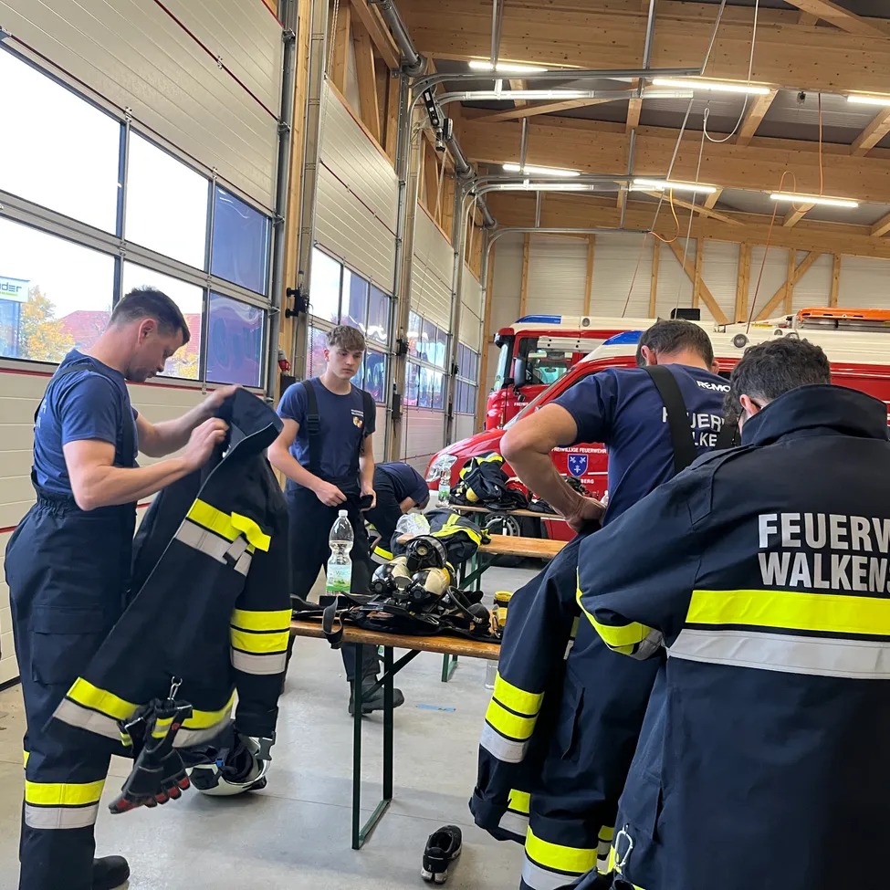 Eine Gruppe von Feuerwehrleuten befindet sich in einer Feuerwache und bereitet ihre Ausrüstung vor. Sie tragen blaue Uniformen mit gelben und weißen Streifen.