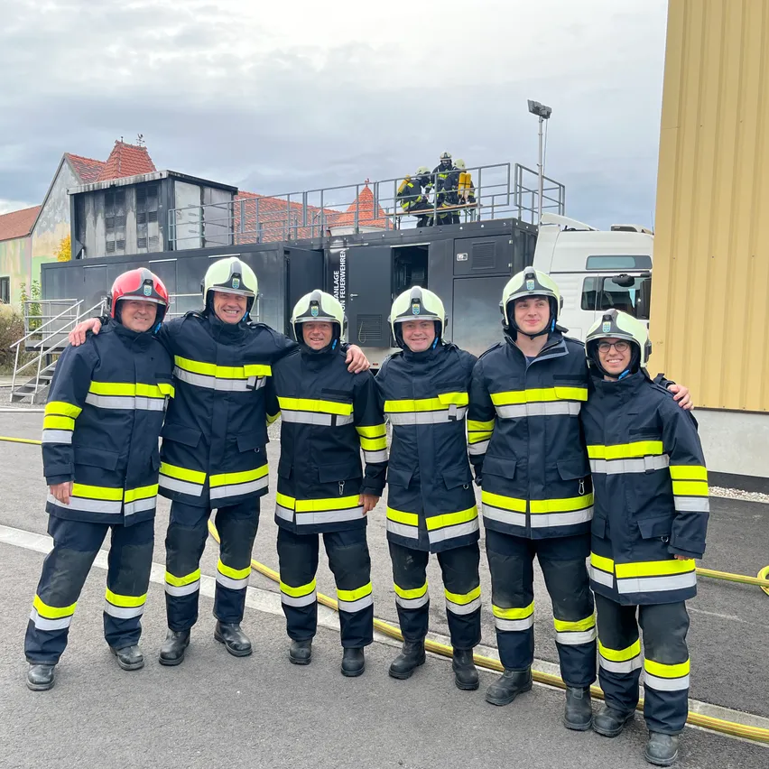 Fünf Feuerwehrleute in voller Ausrüstung stehen vor einem Gebäude mit einem Feuerwehrwagen und einem Feuerwehrschlauch auf dem Boden.