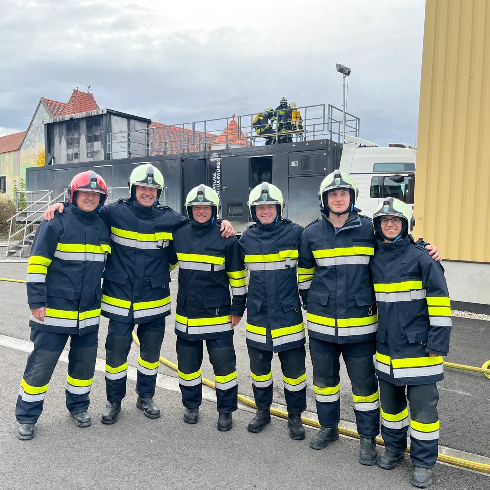 Fünf Feuerwehrleute in voller Ausrüstung stehen vor einem Gebäude mit einem Feuerwehrwagen und einem Feuerwehrschlauch auf dem Boden.