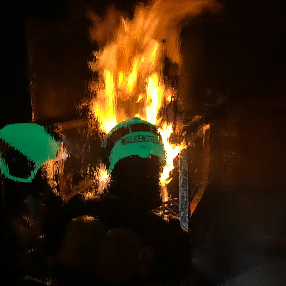 Ein Feuerwehrmann in einem grünen Helm mit dem Wort 'WALKENSTED' darauf steht vor einem Feuer.