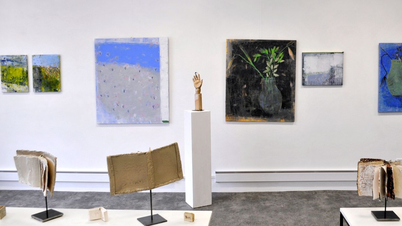 Eine Kunstgalerie zeigt drei Gemälde an weißen Wänden. Eine Skulptur ruht auf einem weißen Podest zwischen zwei Gemälden. Das linke Gemälde hat einen blauen Himmel und einen grauen Boden. Das rechte Gemälde zeigt eine Vase mit Pflanzen. Eine kleine Skulptur und andere Objekte befinden sich auf dem Boden.