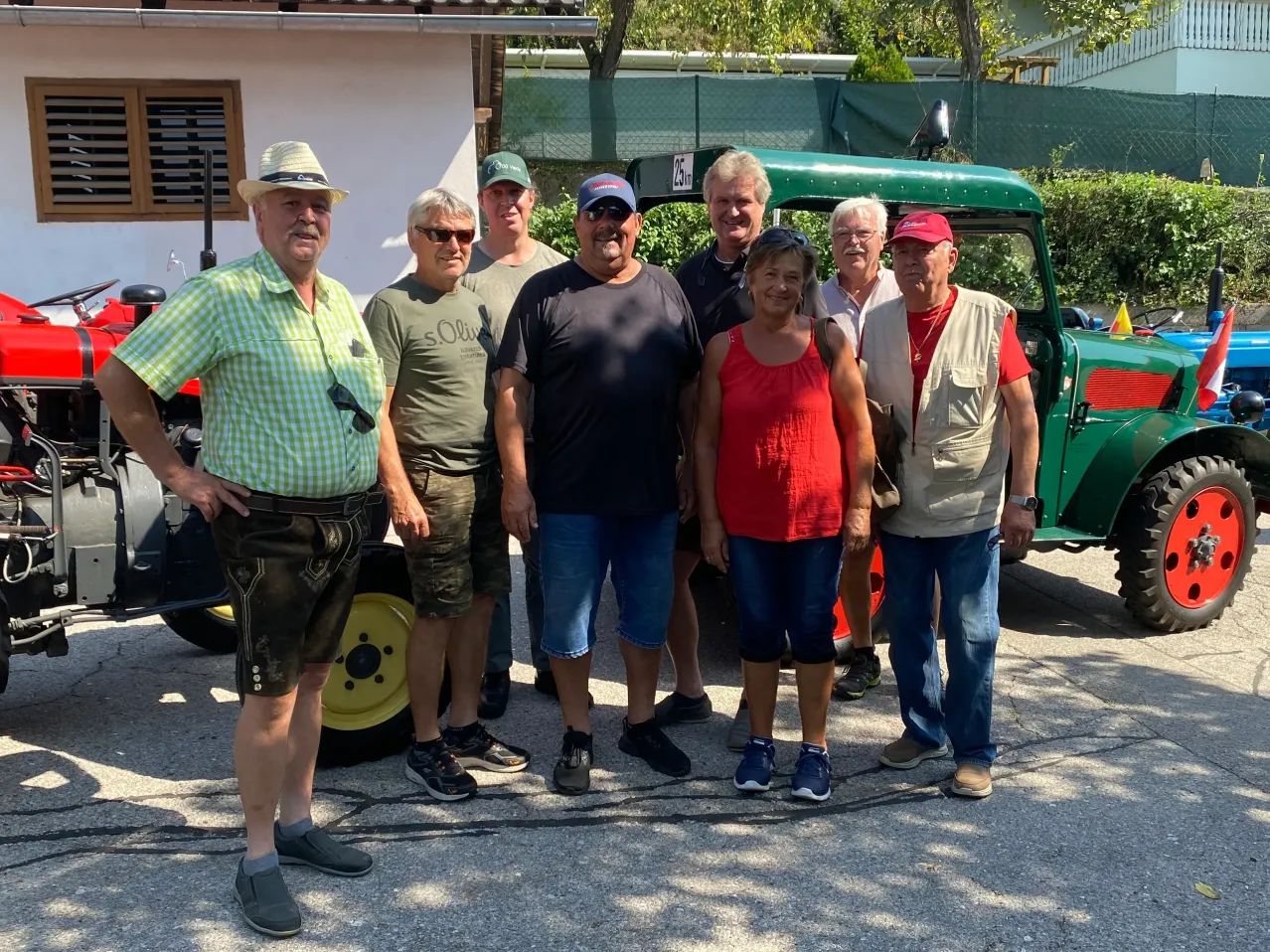 Eine Gruppe von sechs Personen steht vor einem grünen Traktor, lässig gekleidet, mit einem Gebäude und Grünpflanzen im Hintergrund.