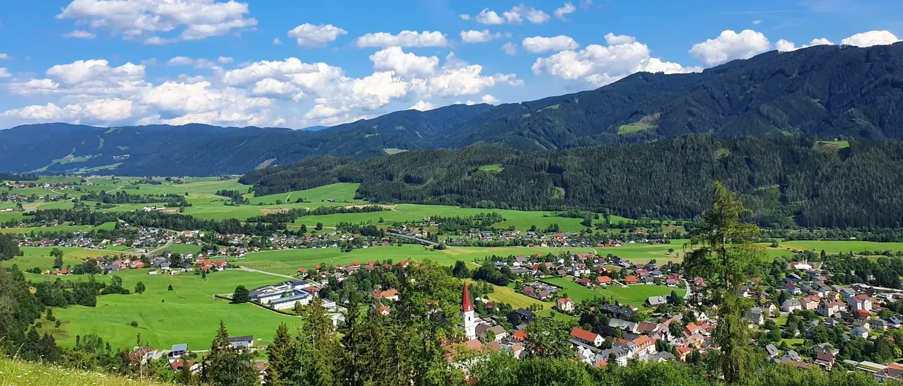 Luftaufnahme eines Dorfes in einem Tal, umgeben von üppigen grünen Feldern und Bergen unter einem teilweise bewölkten Himmel.