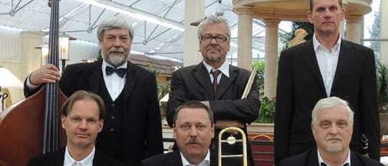 Eine Gruppe von Musikern, die Anzüge und Krawatten tragen, posiert für ein Foto. Der Mann auf der linken Seite hält eine Trompete. Der Mann in der Mitte hält ein Posaune. Der Mann auf der rechten Seite hält eine Gitarre. Dahinter steht ein Pavillon.