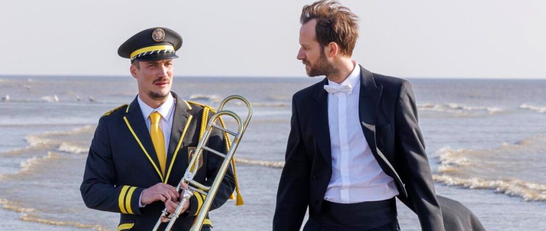 Zwei Männer stehen am Strand. Einer in Uniform hält eine Posaune, der andere in formaler Kleidung unterhält sich. Der Meereshintergrund ist zu sehen.