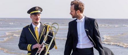 Zwei Männer stehen am Strand. Einer in Uniform hält eine Posaune, der andere in formaler Kleidung unterhält sich. Der Meereshintergrund ist zu sehen.