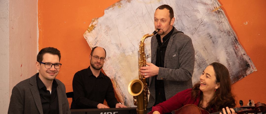 Ein Quartett von Musikern spielt in einem Raum mit einer orangefarbenen Wand. Ein Mann spielt die Saxophon, während ein anderer das Klavier spielt. Eine Frau spielt die Geige, und ein anderer Mann spielt die Trommeln.