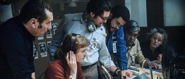 Eine Gruppe von Menschen versammelt sich in einem Raum um einen Schreibtisch, trägt Kopfhörer und schaut auf einen Computerbildschirm. Die Frau in der Mitte hält ein Mikrofon.