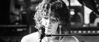 Ein Schwarz-Weiß-Foto einer Frau, die Klavier spielt. Sie trägt einen weißen Pullover und hat lockige Haare. Sie singt in ein Mikrofon.