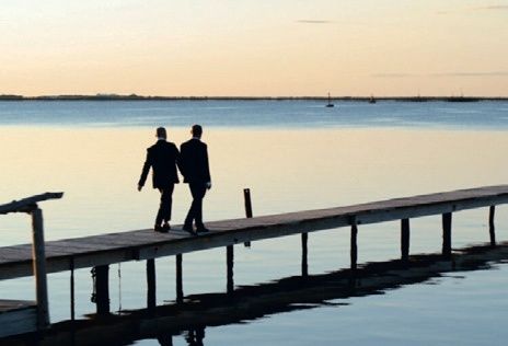 Zwei Männer in Anzügen gehen auf einem hölzernen Steg über eine ruhige Wasserfläche bei Sonnenuntergang.