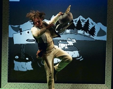 Eine Person springt auf einer Bühne mit einem Hintergrund einer verschneiten Landschaft und Bäumen. Die Person hält einen Hut in beiden Händen.