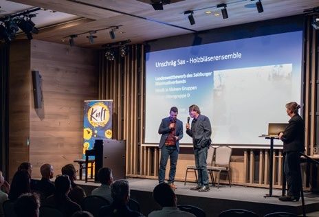 Drei Männer stehen auf einer Bühne vor einem Publikum mit einer Projektionsleinwand im Hintergrund, die 'Unschrlig Sax Holzblaserensemble' anzeigt.