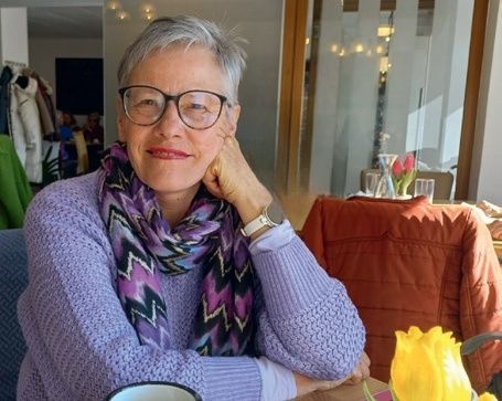 Eine Frau mit Brille und Schal sitzt an einem Tisch mit einer Tasse und einer gelben Blume in einer Vase.