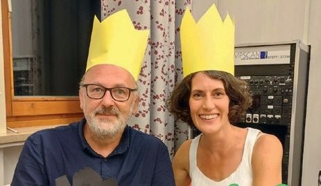 Ein älterer Mann und eine Frau tragen gelbe Papierkronen und lächeln für ein Foto. Der Mann hat einen Bart und eine Brille. Sie stehen vor einem Vorhang und einem Rack mit einer Markenbezeichnung.