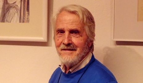 Ein älterer Mann mit weißen Haaren und Bart steht vor einer Wand und trägt einen blauen Pullover. Dahinter hängen zwei gerahmte Bilder an der Wand.