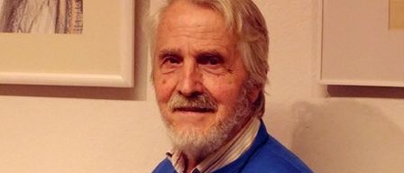 Ein älterer Mann mit weißen Haaren und Bart steht vor einer Wand und trägt einen blauen Pullover. Dahinter hängen zwei gerahmte Bilder an der Wand.