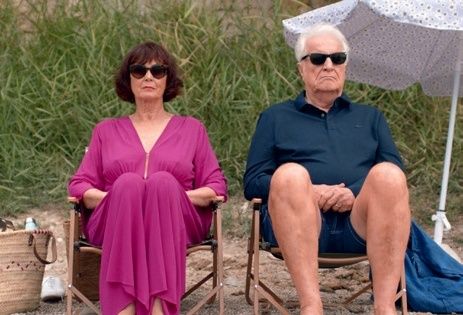 Zwei ältere Erwachsene sitzen auf Stühlen am Strand, eine in einem lila Kleid und der andere in einem blauen Hemd. Sie haben einen Regenschirm hinter sich und einen Korb in der Nähe der Frau.