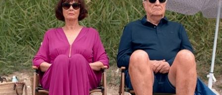 Zwei ältere Erwachsene sitzen auf Stühlen am Strand, eine in einem lila Kleid und der andere in einem blauen Hemd. Sie haben einen Regenschirm hinter sich und einen Korb in der Nähe der Frau.