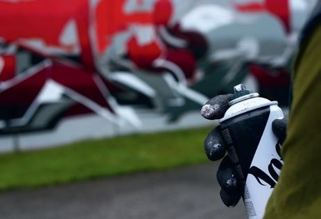 Eine Person hält eine Spraydose mit Graffiti an der Wand hinter ihr. Die Graffiti sind überwiegend rot und grau.