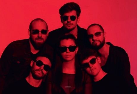 Eine Gruppe von Musikern, fünf Männer und eine Frau, mit Sonnenbrillen posiert für ein Foto vor einem roten Hintergrund.