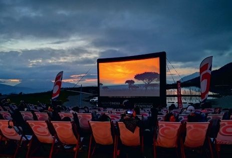 Ein Open-Air-Kino mit einer großen Leinwand, die einen Sonnenuntergang zeigt. Die Menschen sitzen in Stuhlreihen, einige mit Fahnen dahinter. Berge und ein Gewässer sind in der Ferne.