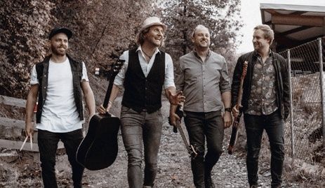 Vier Männer, Gitarren in der Hand, gehen auf einem von Bäumen und einem Zaun umgebenen Weg. Sie sehen glücklich aus und sind in verschiedenen lässigen Outfits gekleidet.