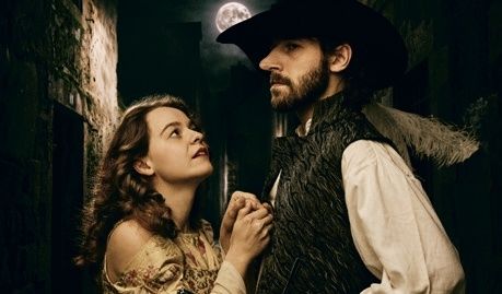 Ein Mann und eine Frau, gekleidet in historischer Kleidung, stehen eng in einer dunklen Gasse, wobei ein Vollmond die Szene hinter ihnen beleuchtet.