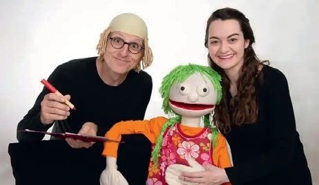 Zwei Personen sitzen mit einer Puppe, eine hält einen roten Marker und die andere hält eine Puppe mit grünen Haaren. Sie tragen beide schwarze Langarmshirts.