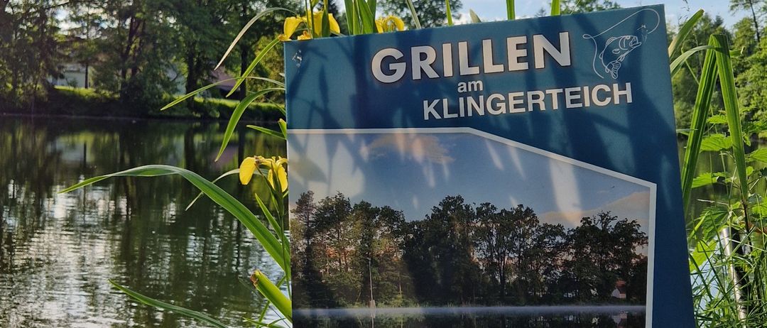 Ein blaues Schild steht am See und bewirbt 'Grillen am Klinger Teich' mit einem Bild des Sees, der Bäume und der Reflexionen. Es ist für eine Veranstaltung am 15. Juni ab 10 Uhr.