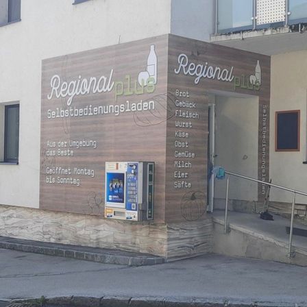 Ein Ladenausbau mit einem deutschen Schild, das Produkte wie Brot, Käse, Fleisch, Wurst, Käse, Milch, Gemüse, Eier und Säfte anbietet. Ein Zahlungsautomat ist an der Wand montiert.