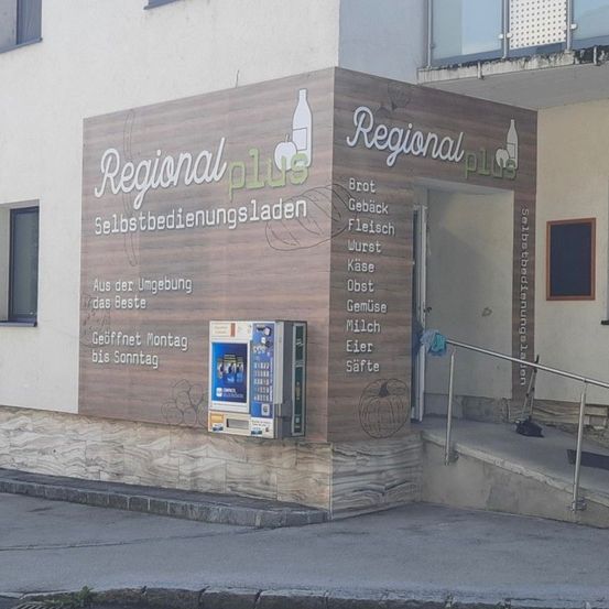 Ein Ladenausbau mit einem deutschen Schild, das Produkte wie Brot, Käse, Fleisch, Wurst, Käse, Milch, Gemüse, Eier und Säfte anbietet. Ein Zahlungsautomat ist an der Wand montiert.