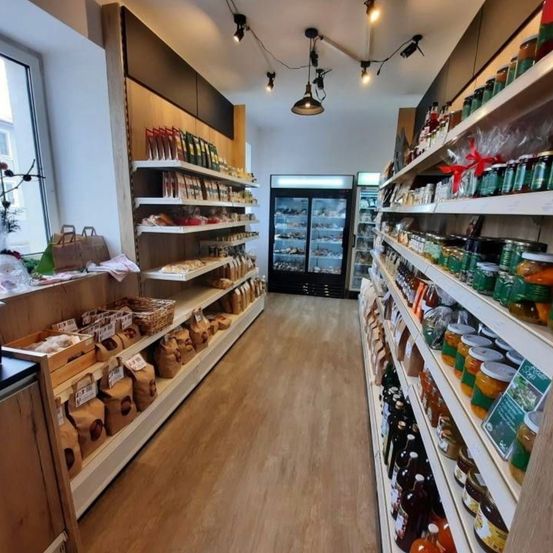 Ein Gang in einem Lebensmittelgeschäft mit Holzregalen, die mit verschiedenen verpackten Lebensmitteln gefüllt sind, darunter Brot, Kekse und Konserven, sowie einem Kühlschrank im Hintergrund.