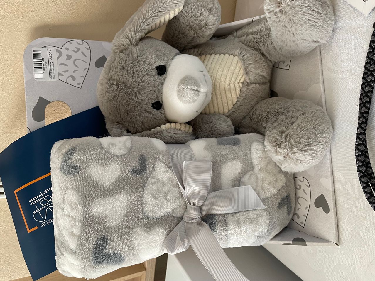 Ein grauer Plüschtier mit einer weißen Nase und einer weißen Schleife liegt auf einer grauen Decke in einer weißen Schachtel. Die Schachtel hat einen blauen Deckel mit einem Herzdesign.