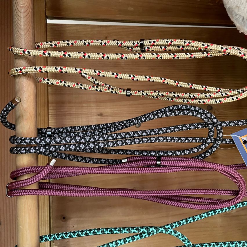 Eine hölzerne Oberfläche zeigt fünf Seile in verschiedenen Farben und Mustern. Jedes Seil hat einen Metallring an einem Ende. Die Seile sind beige, schwarz, braun und türkis.