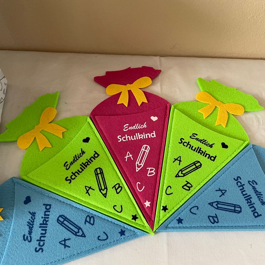 Eine bunte Anordnung von dreieckigen Lesezeichen, jeweils in verschiedenen Farben mit dem Wort 'Endlich Schulkind' und den Buchstaben A, B, C, D und E, auf einem weißen Tuchhintergrund.