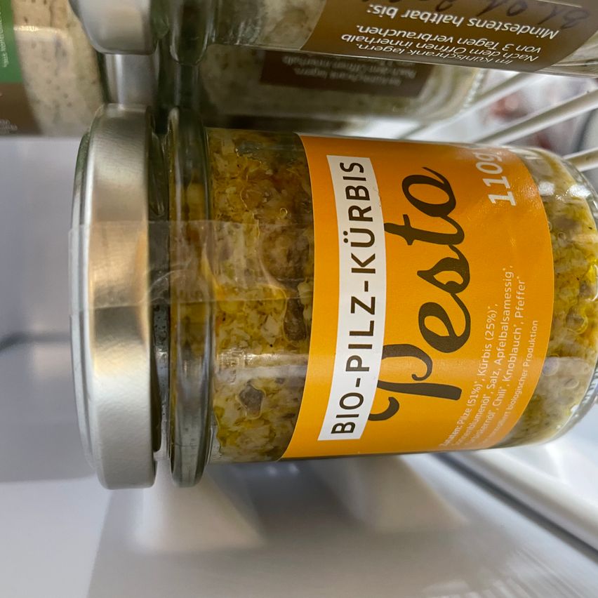 Ein Glasgefäß mit silbernem Deckel, gefüllt mit gelben Zutaten, beschriftet mit 'Bio-Pflanz-Kurbis Pesto'. Weitere Gläser sind im Hintergrund sichtbar.