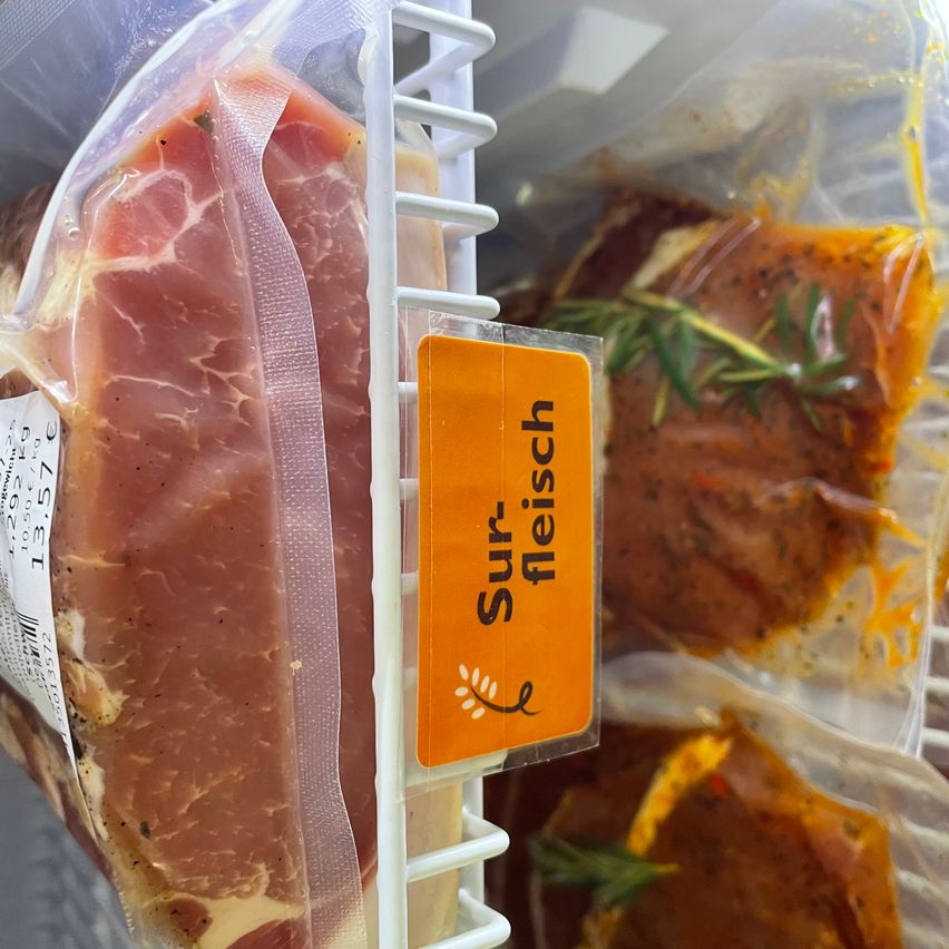 Ein verpacktes Stück Fleisch mit einem Schild, das 'Surfleisch' liest, befindet sich auf einem weißen Regal mit anderen Artikeln in einem Kühlschrank.